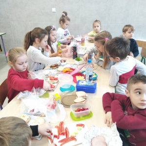 Uczniowie z „zerówki” i klasy I wspólnie przy długim stole przygotowują kolorowe kanapki.