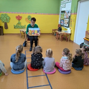 Zdjęcie przedstawia nauczyciela-bibliotekarza pokazującego ilustracje z czytanej książki dzieciom z przedszkola