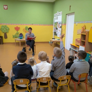 Zdjęcie przedstawia nauczyciela-bibliotekarza rozmawiającego z dziećmi z przedszkola i z oddziału przedszkolnego w ramach cyklicznej akcji czytania bajek w przedszkolu.